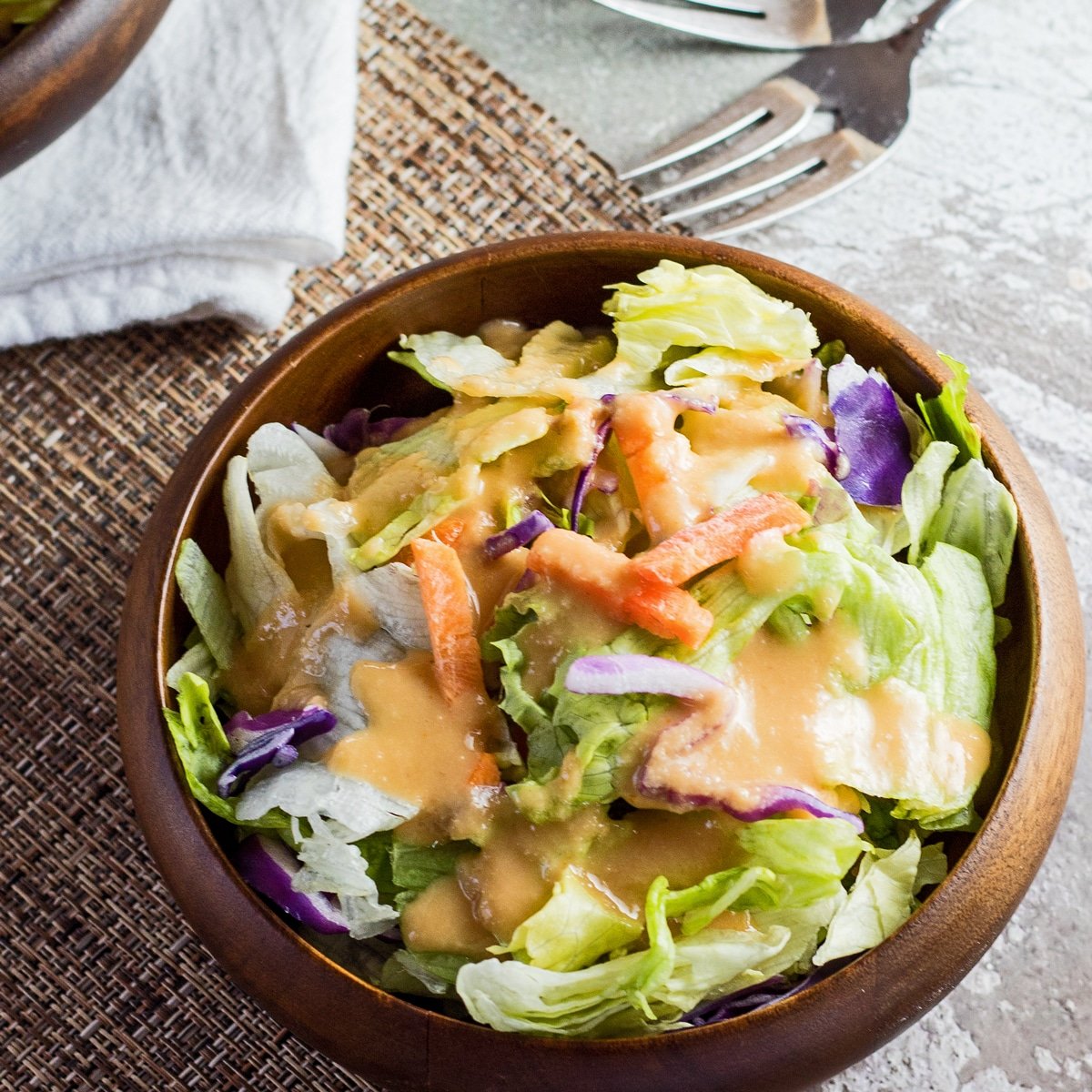 Stort firkantet billede af benihana ingefær salatdressing serveret over salat i en mørk bambusskål med lys tekstureret baggrund.