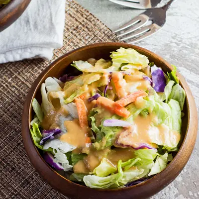 small square image of the benihana ginger salad dressing served over salad in a dark bamboo bowl with light textured background