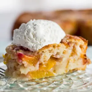 large square image closeu sideview of baked nectarine cobbler sliced and served with a scoop of whipped cream on a crystal plate with the cobbler in background