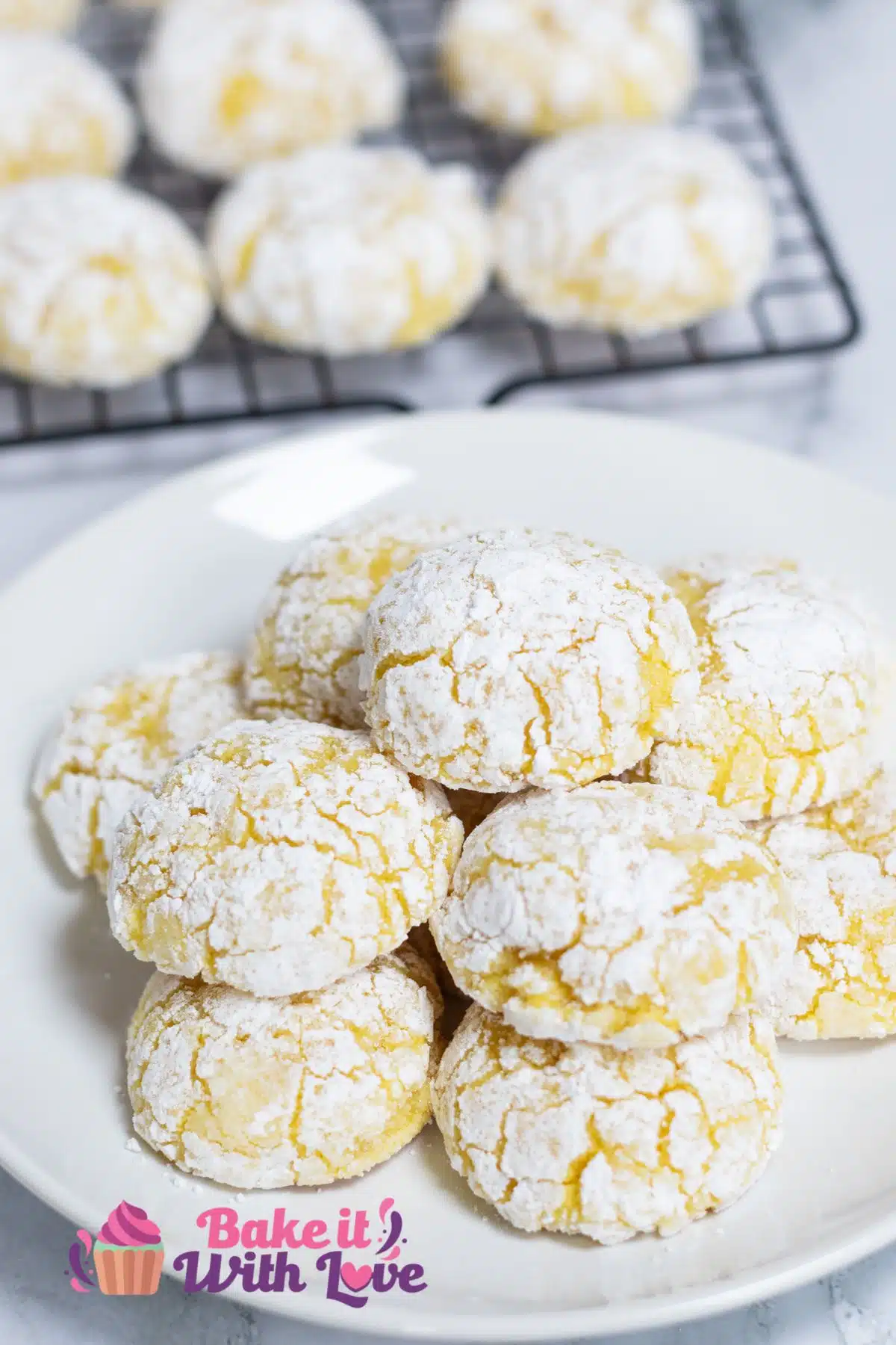 Best lemon cream cheese crinkle cookies recipe as shown with cookies piled on a white plate and more in the background on wire cooling rack.