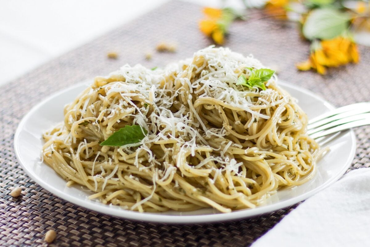large horizontal image angled side view of dished pasta with pesto coating garnished with fresh grated Parmesan cheese and a few young Genovese basil leaves
