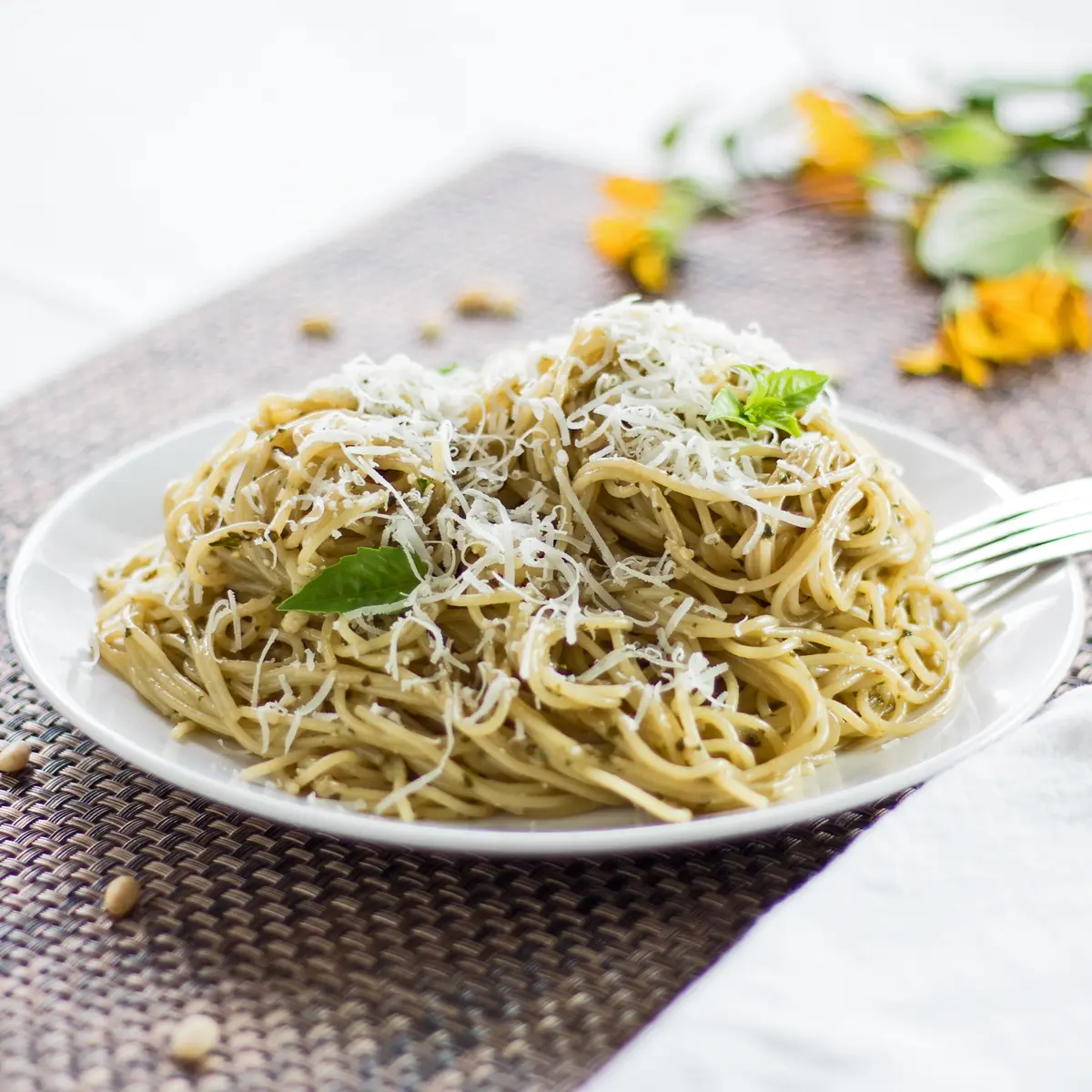 Large square image angled side view of dished pasta with pesto coating garnished with fresh grated Parmesan cheese and a few young Genovese basil leaves.