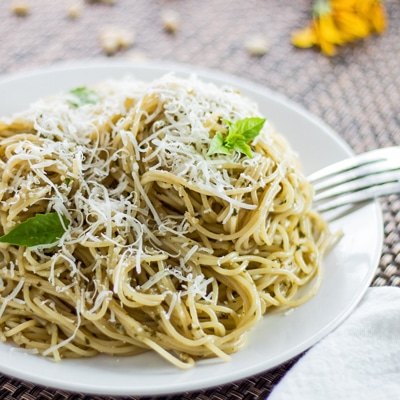 small square cropped closeup angled overhead of dished pasta with pesto coating garnished with fresh grated Parmesan cheese and a few young Genovese basil leaves