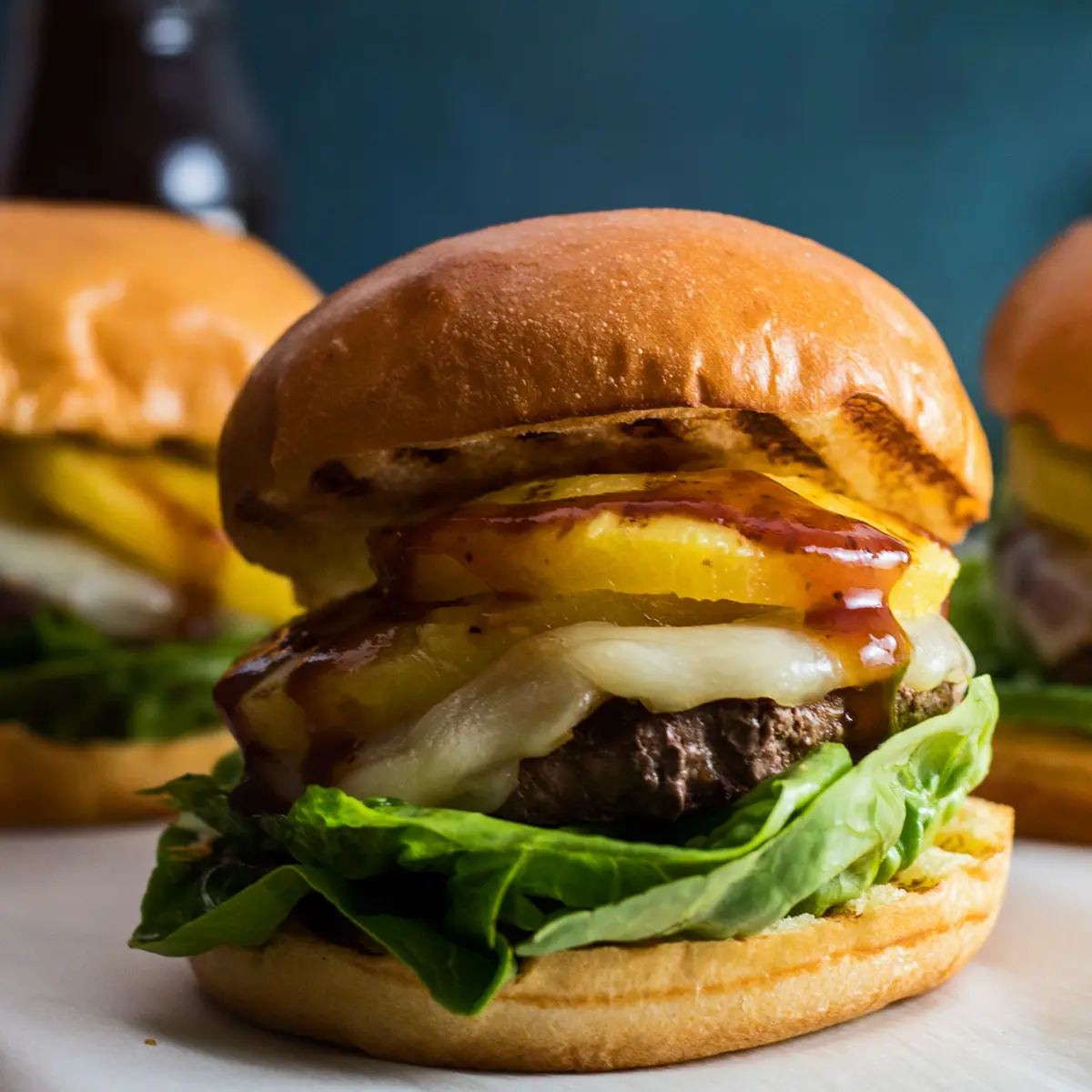 Large square image straight on closeup of the stacked Hawaiian Burger ingredients including ground beef patty, provolone cheese, grilled pineapple rings, lettuce, and teriyaki sauce on grilled brioche buns.