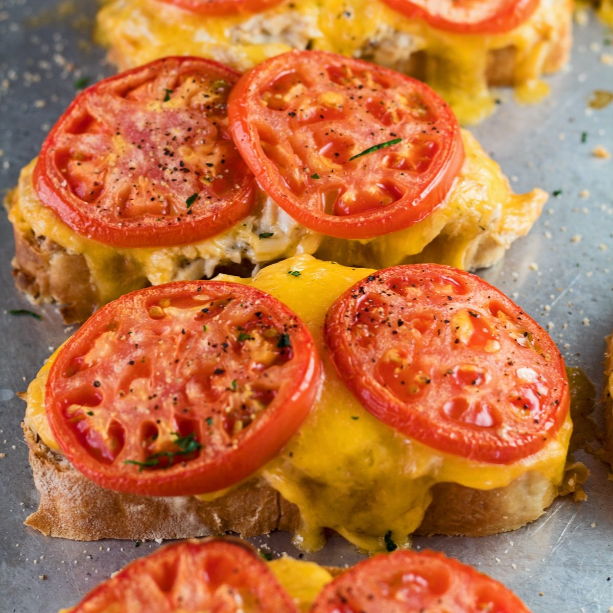 Large square angled overhead image of tuna melt sanwiches still open faced after broiling to heat the tuna salad, cheddar cheese and sliced tomatoes on the baking sheet.