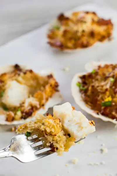 vertical image closeup on a cut away bite of the scallop on a fork with other breaded stuffed scallops in shells in the background