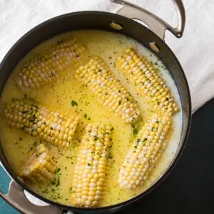 Halved sweet corn cobs shown in the water, milk, and butter mixture that they were boiled in on the stovetop.