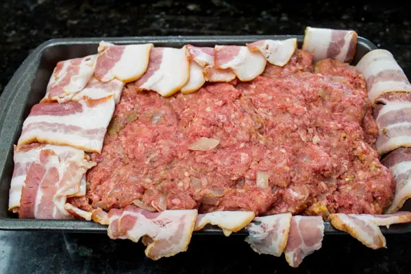 bacon wrapped meatloaf being formed in the loaf pan with the edges of bacon being tucked back under