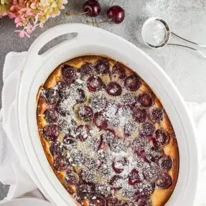 large square overhead image of the baked bing cherry clafoutis in the white oval baking dish with cherries scattered around on a light grey background