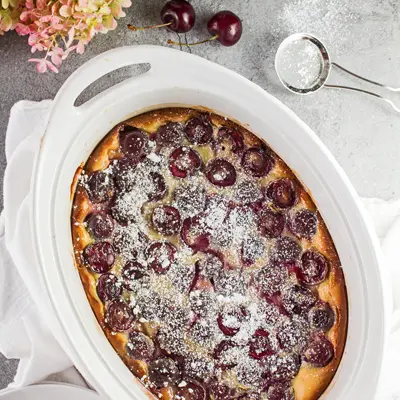 small square overhead image of the baked bing cherry clafoutis in the white oval baking dish with cherries scattered around on a light grey background