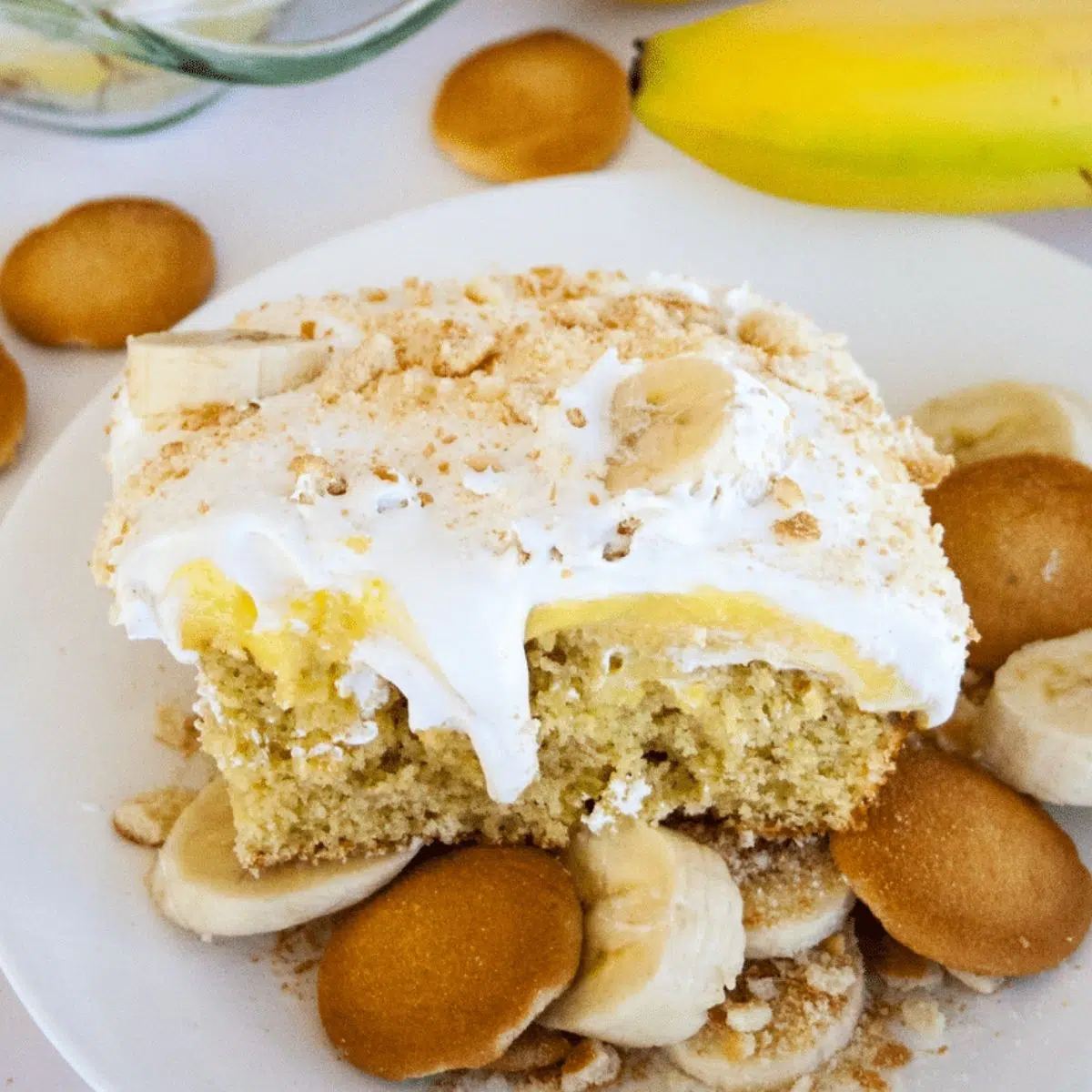 Delicious slice of banana pudding poke cake served with more bananas and Nilla wafers on white plate.