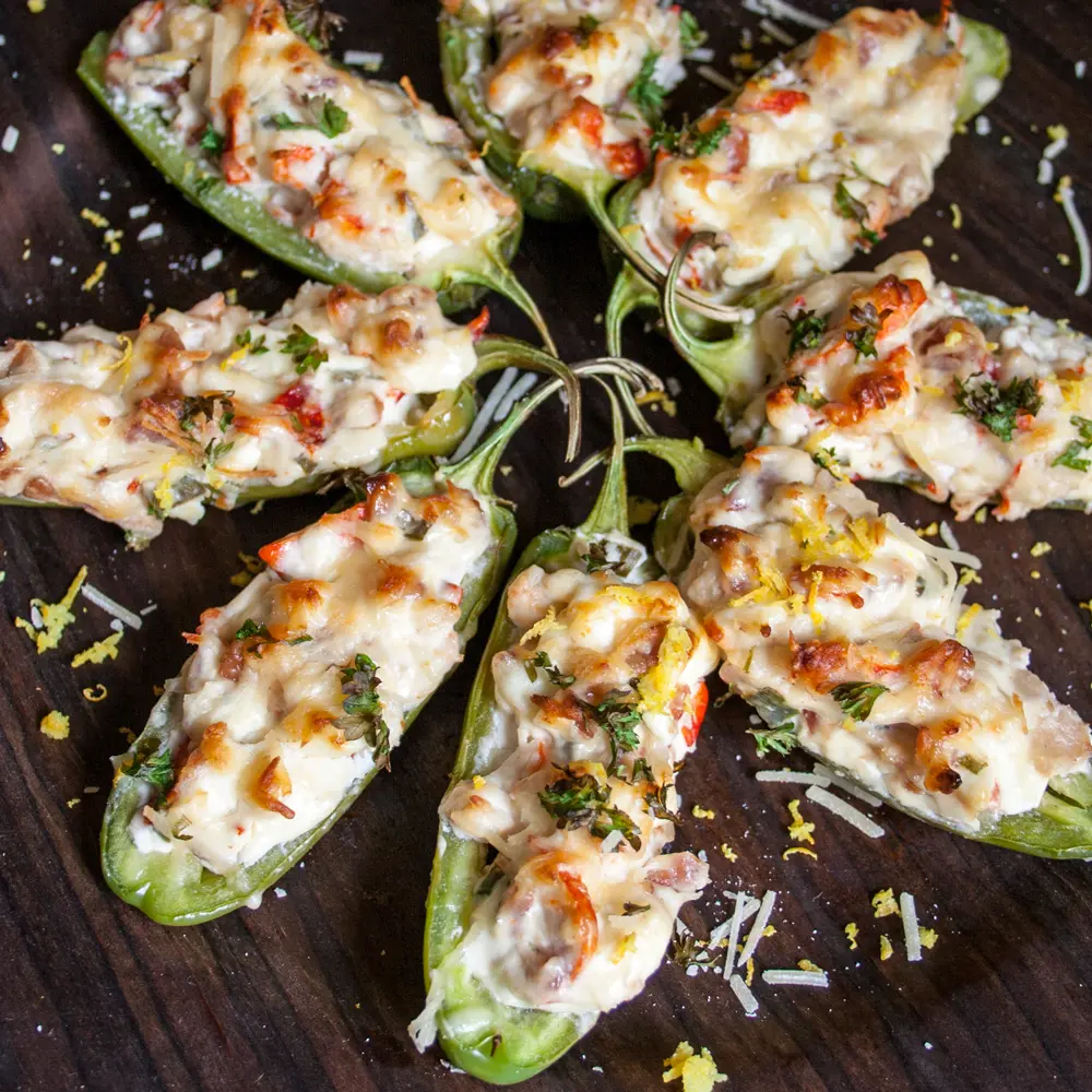 Top-down view of eight lobster jalapeño poppers displayed in a starburst pattern on a dark wooden background. Each jalapeño half is filled with a rich lobster and cheese mixture, melted and golden on top, and garnished with chopped herbs, Parmesan, and lemon zest.