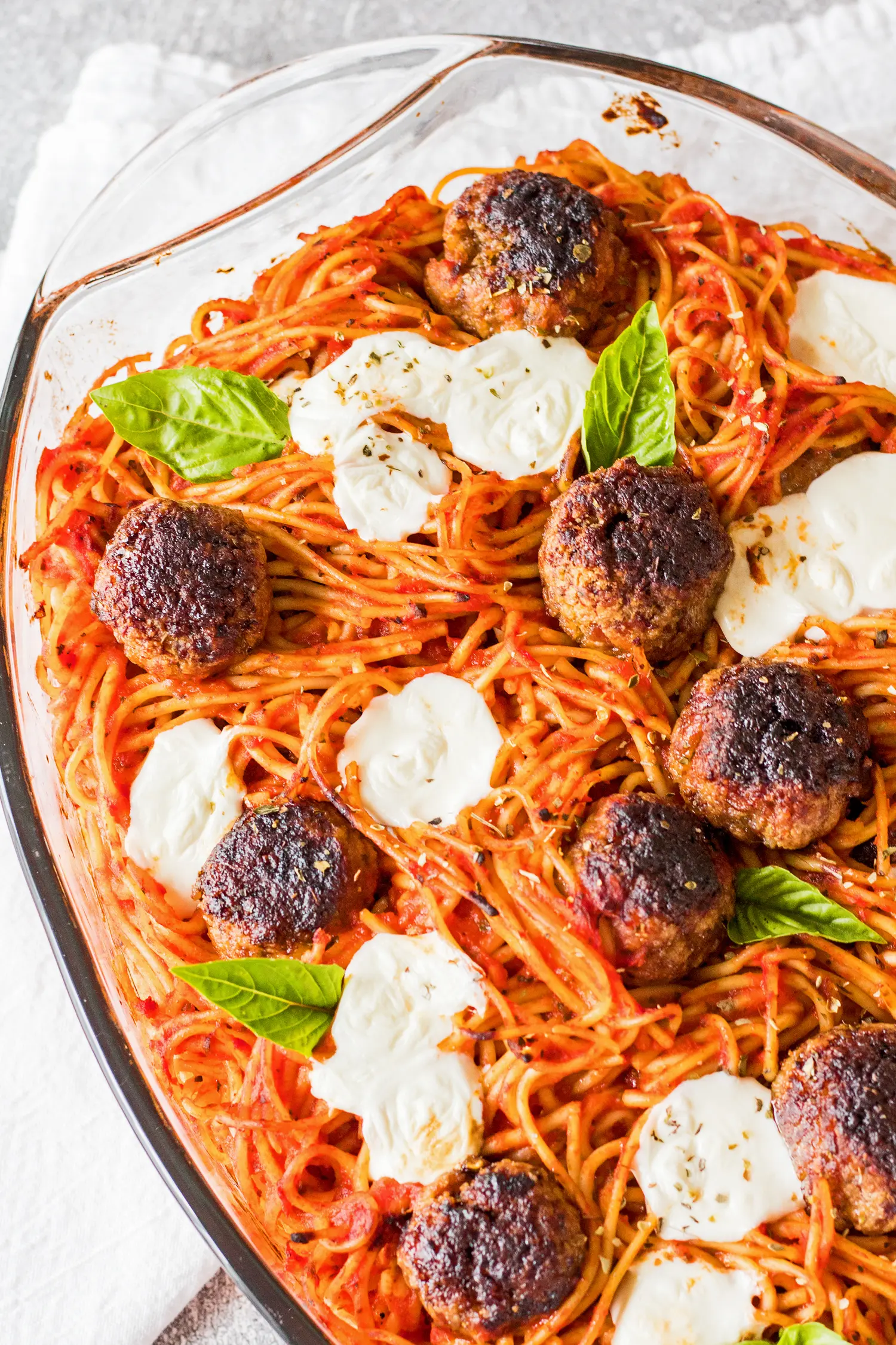 Overhead vertical image of baked spaghetti and meatballs in a clear casserole dish baked with fresh baby mozzarella and garnished with genovese basil leaves on a light grey background.