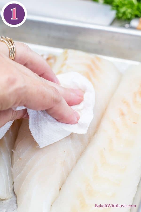 Lemon butter baked cod process photo 1. A hand with rings is seen gently dabbing a white fish fillet with a folded white paper towel, preparing it on a silver baking pan. Other unpatched fillets are also present on the tray. A purple circle with the number "1" is in the upper left corner.