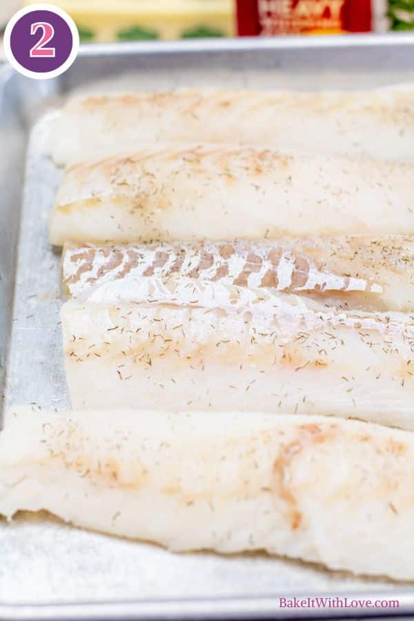 Lemon butter baked cod process photo 2. Several pieces of raw, white fish fillets, lightly seasoned with dark green specks of herbs, are arranged on a silver baking sheet. One fillet shows a hint of reddish-brown skin. A purple circle with the number "2" is in the top left corner.