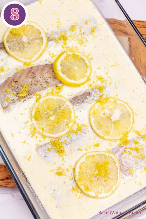 Lemon butter baked cod process photo 8. An overhead view captures a glass baking dish filled with milky sauce, fish fillets, and vibrant yellow lemon slices. Sprinkled over the dish are fine lemon zest and dark green herbs. A purple circle containing the number "8" is visible in the top left.