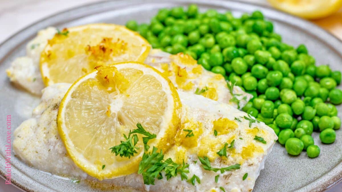 A close-up, horizontal shot of two filets of lemon butter baked cod on a light grey plate. Each white fish fillet is topped with a thin slice of baked lemon and garnished with fresh parsley. To the right of the cod, there's a pile of bright green peas. The plate sits on a blurred background, possibly a light-colored table or counter. The text "BakeItWithLove.com" is visible vertically along the left edge.