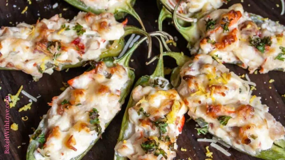 A wide image showing a top-down view of eight lobster jalapeño poppers displayed in a starburst pattern on a dark wooden background. Each jalapeño half is filled with a rich lobster and cheese mixture, melted and golden on top, and garnished with chopped herbs, Parmesan, and lemon zest.
