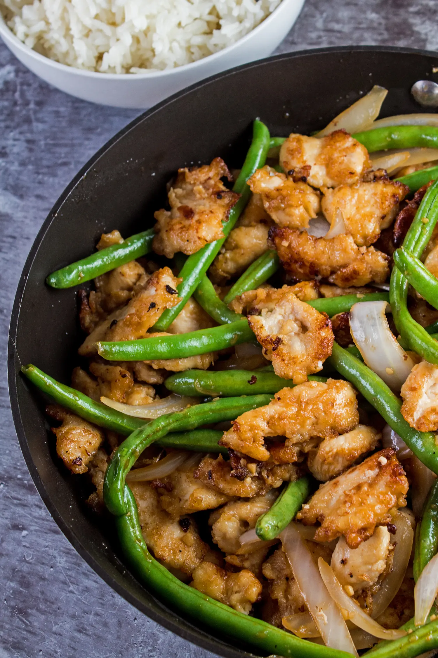 Tall vertical overhead image of the panda express string bean chicken with green beans, chunks of white onion and chicken breast pieces in a frying pan on grey background.