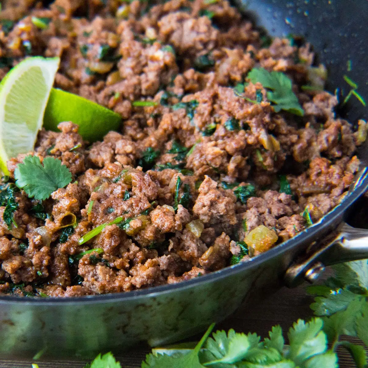 Square image of taco meat in cooked in a pan with a lime and cilantro.