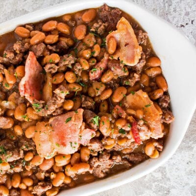Square image of baked beans with bacon in a white casserole dish.