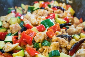 assembled kung pao chicken being wok fried, including bell peppers, zuchhini, chicken chunks, dried peppers and peanuts