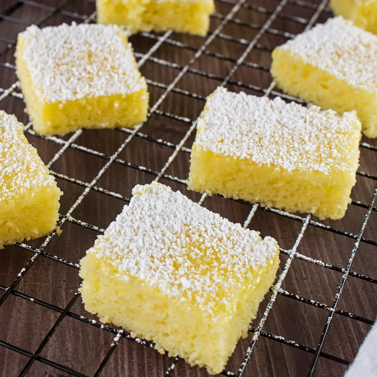 Large square image of easy two ingredient lemon bars cut and dusted with powdered sugar, set on a black wire cooling rack over wood grain surface