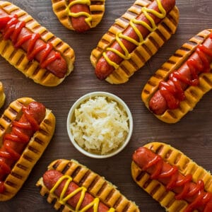 Overhead square image at top showing air fryer hot dogs in buns with ketchup and mustard on brown background with a white bowl of sauerkraut near the center.