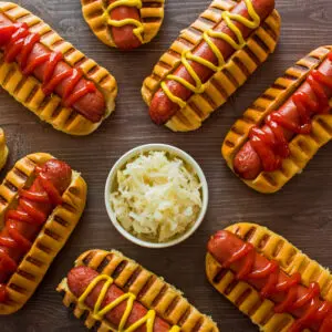 Overhead square image at top showing air fryer hot dogs in buns with ketchup and mustard on brown background with a white bowl of sauerkraut near the center.