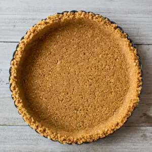 square overhead image of a completed homemade graham cracker crust in a metal fluted round tart pan on a light wooden background