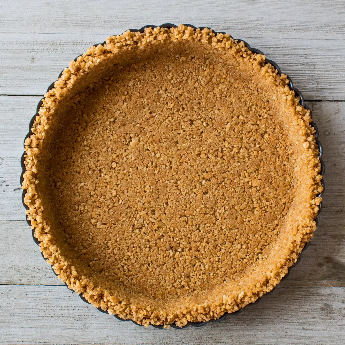 imagem quadrada de cima de uma crosta de biscoito de graham caseiro concluída em uma forma de torta redonda de metal estriada em um fundo claro de madeira