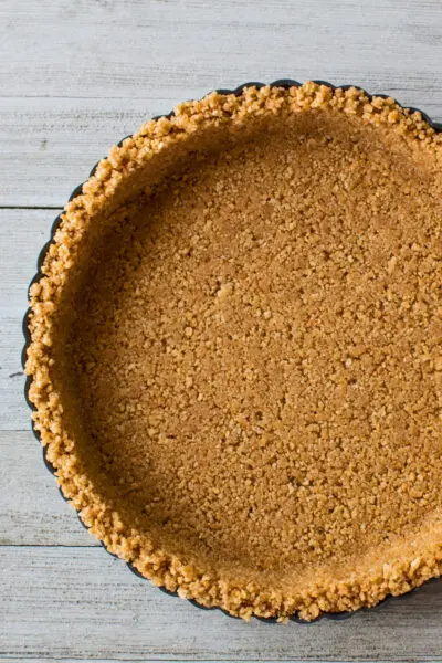 larger vertical overhead image of a completed homemade graham cracker crust in a metal fluted round tart pan on a light wooden background
