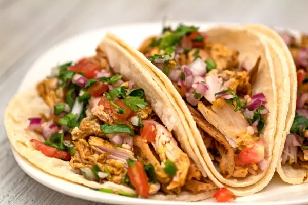 horizontal image showing a close up view of the turkey carnitas garnished and served with fresh homemade pico de gallo served on white corn tortillas on a white plate with light wood grain background