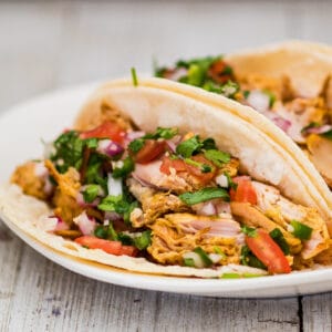 Image carrée montrant une vue rapprochée des carnitas de dinde garnie et servie avec du pico de gallo fait maison frais servi sur des tortillas de maïs blanc sur une plaque blanche avec un fond de grain de bois clair