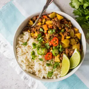 large square overhead image of picodillo mexicano with ground beef tomatoes potatoes vegetables and peppers served with white rice garnished with chopped cilantro and lime wedges on a light teal and white towel with grey background