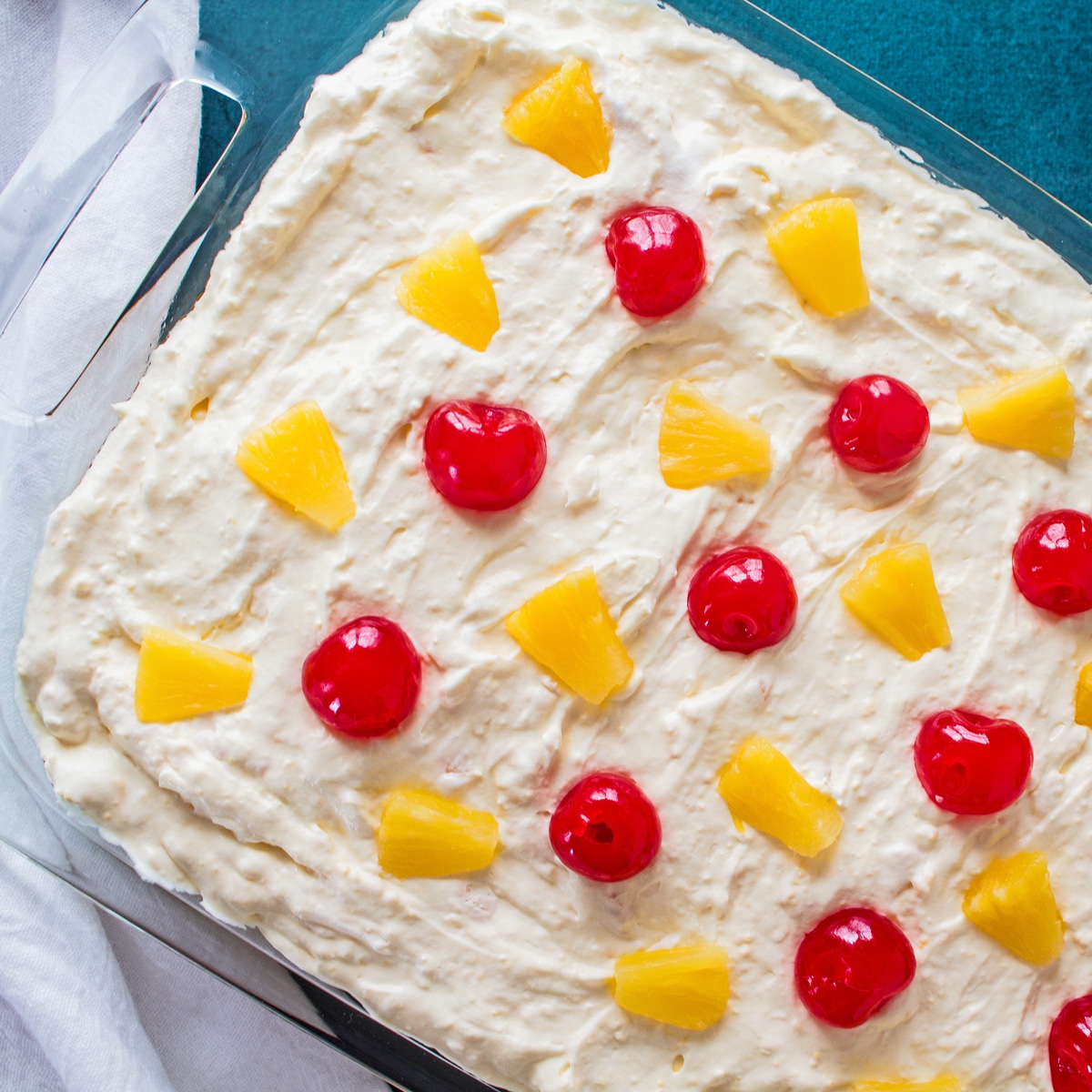 square image showing an overhead of the assembled pineapple sunshine cake in the 9 x 13 pan with green background and garnished with pineapple chunks and maraschino cherries