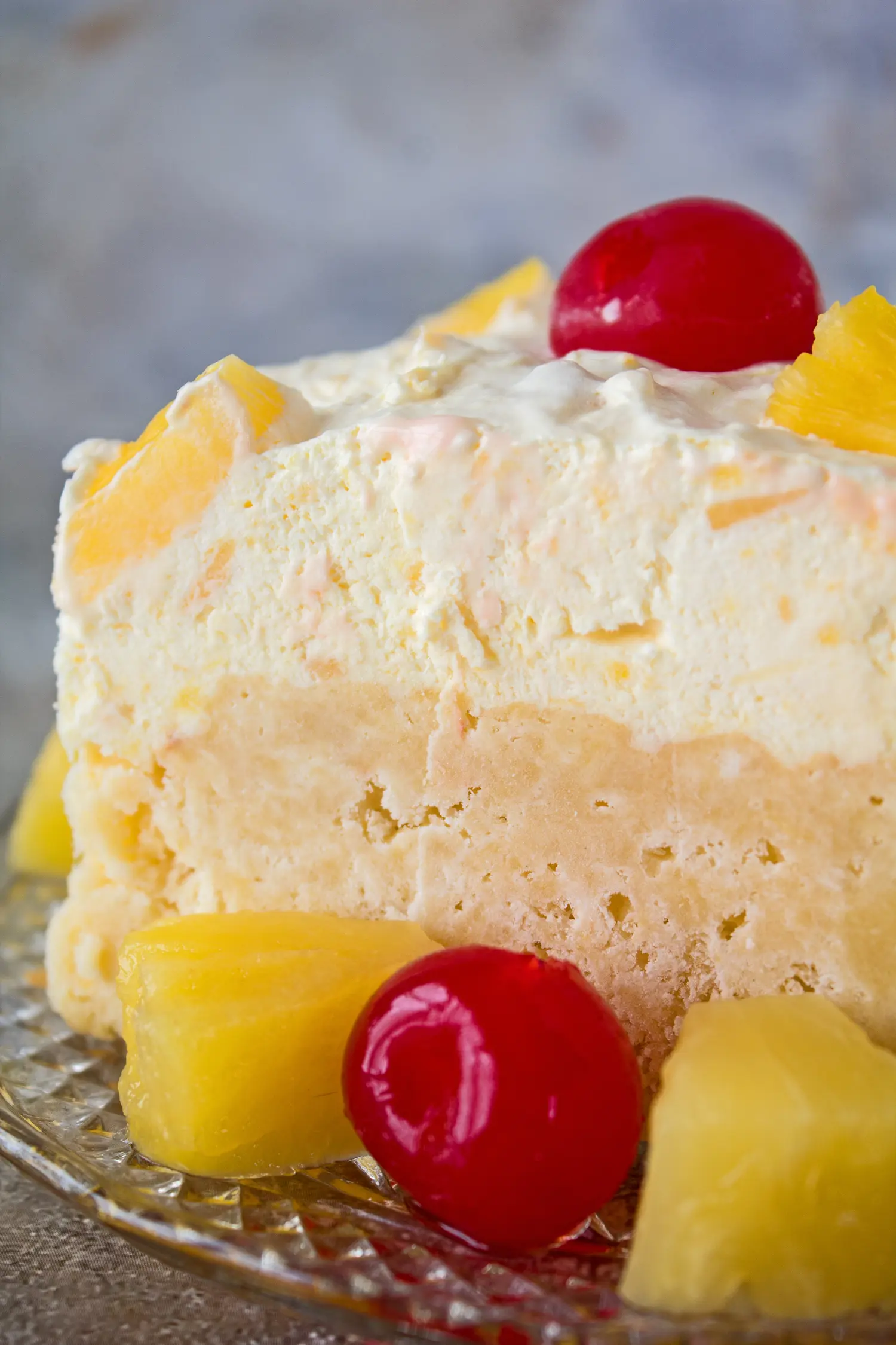 a vertical image of the pineapple sunshine cake sliced and served on a crystal plate with pineapple chunks and maraschino cherries