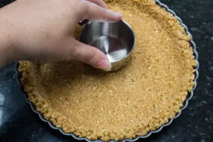 graham cracker crust being tamped down to firmly pack the crust into baking dish