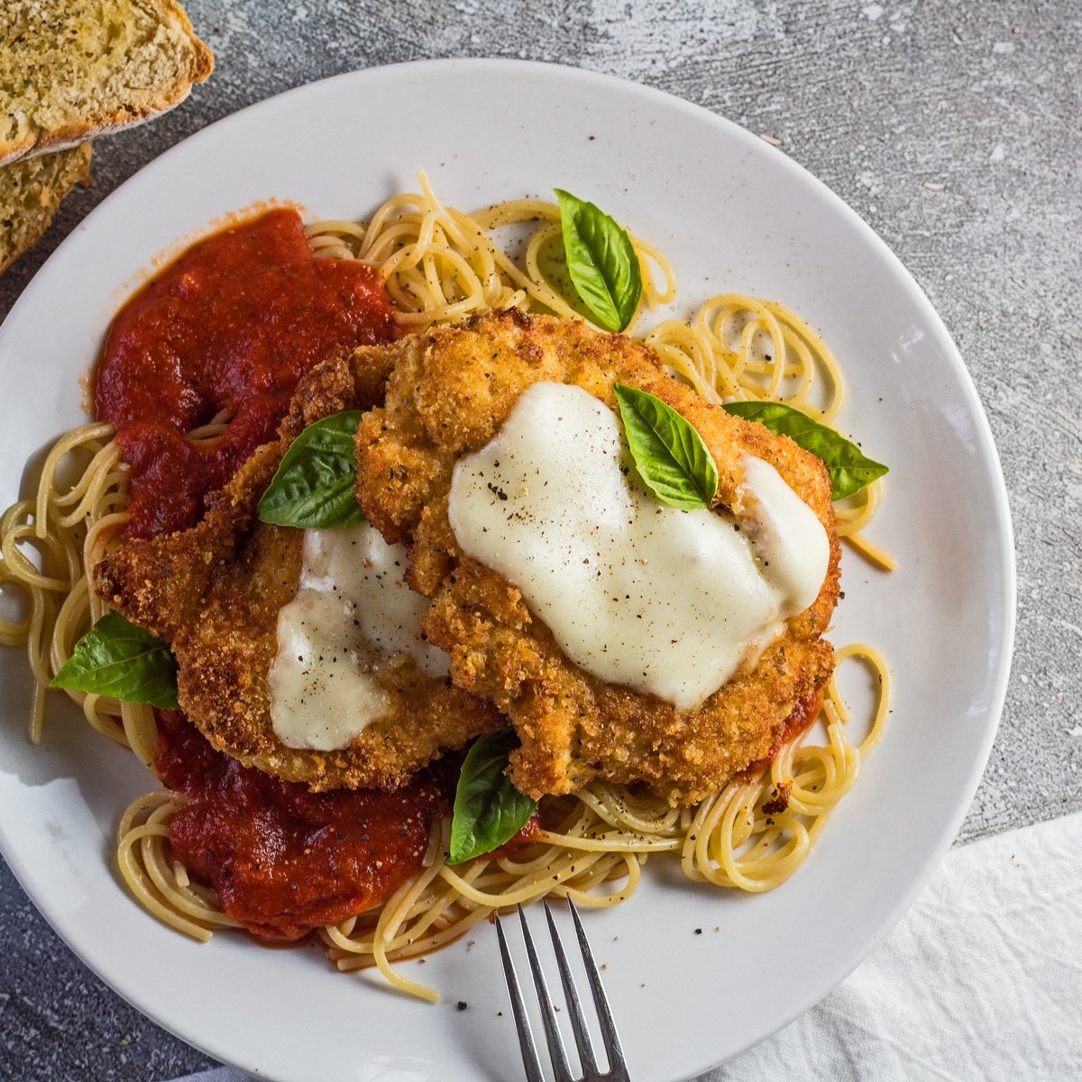 Large square photo showing an overhead view of the chicken parmesan served over a bed of thin spaghetti pasta with marinara sauce and two Panko and Parmesan crusted chicken breast cutlets with melted mozzarella on the center of each cut garnished with a few fresh leaves of baby Genovese basil.
