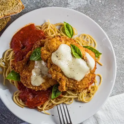 small square photo showing an overhead view of the chicken parmesan served over a bed of thin spaghetti pasta with marinara sauce and two Panko and Parmesan crusted chicken breast cutlets with melted mozzarella on the center of each cut garnished with a few fresh leaves of baby Genovese basil