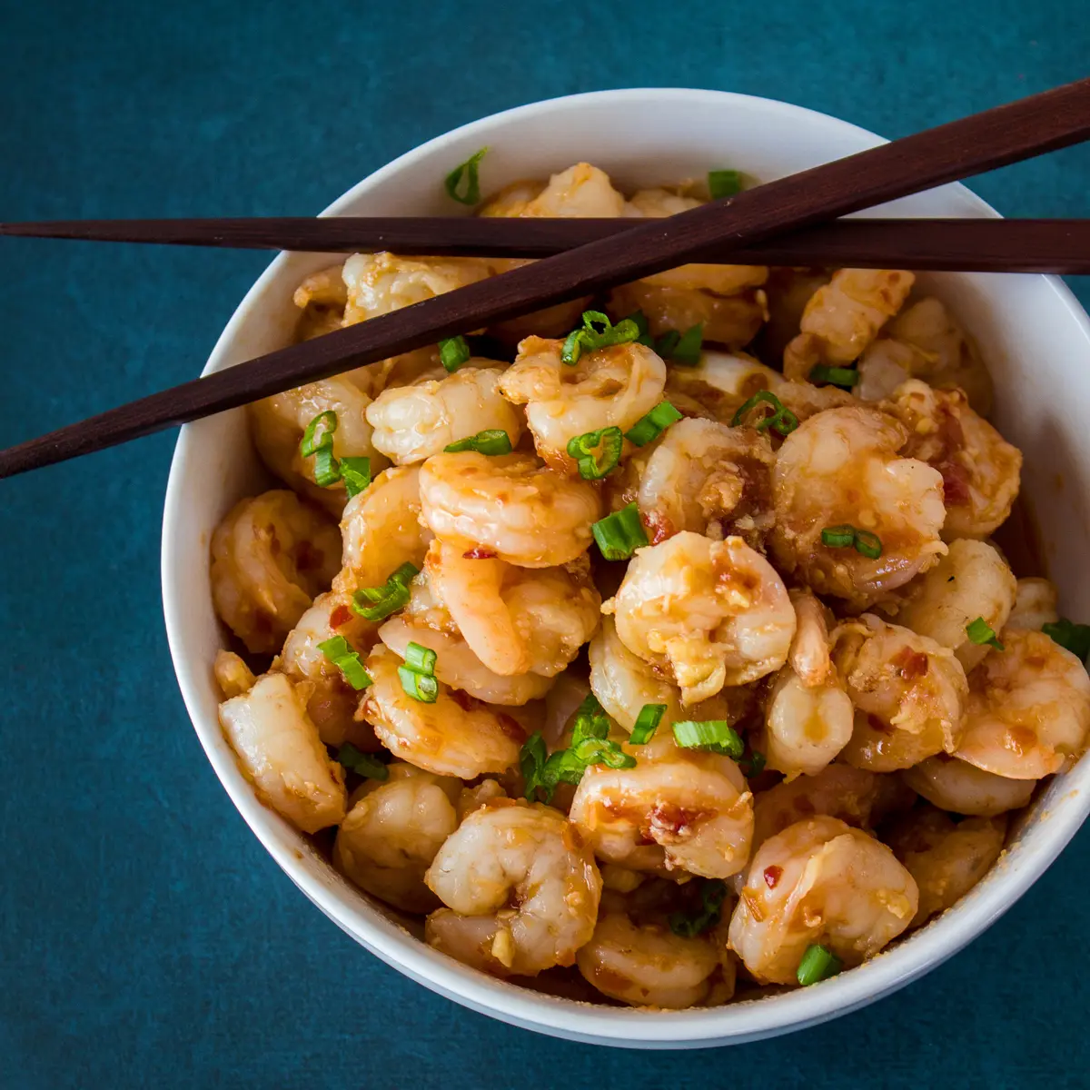 Large square overhead image of hunan shrimp served in a white bowl with chopsticks placed on rim set on a dark blue background.