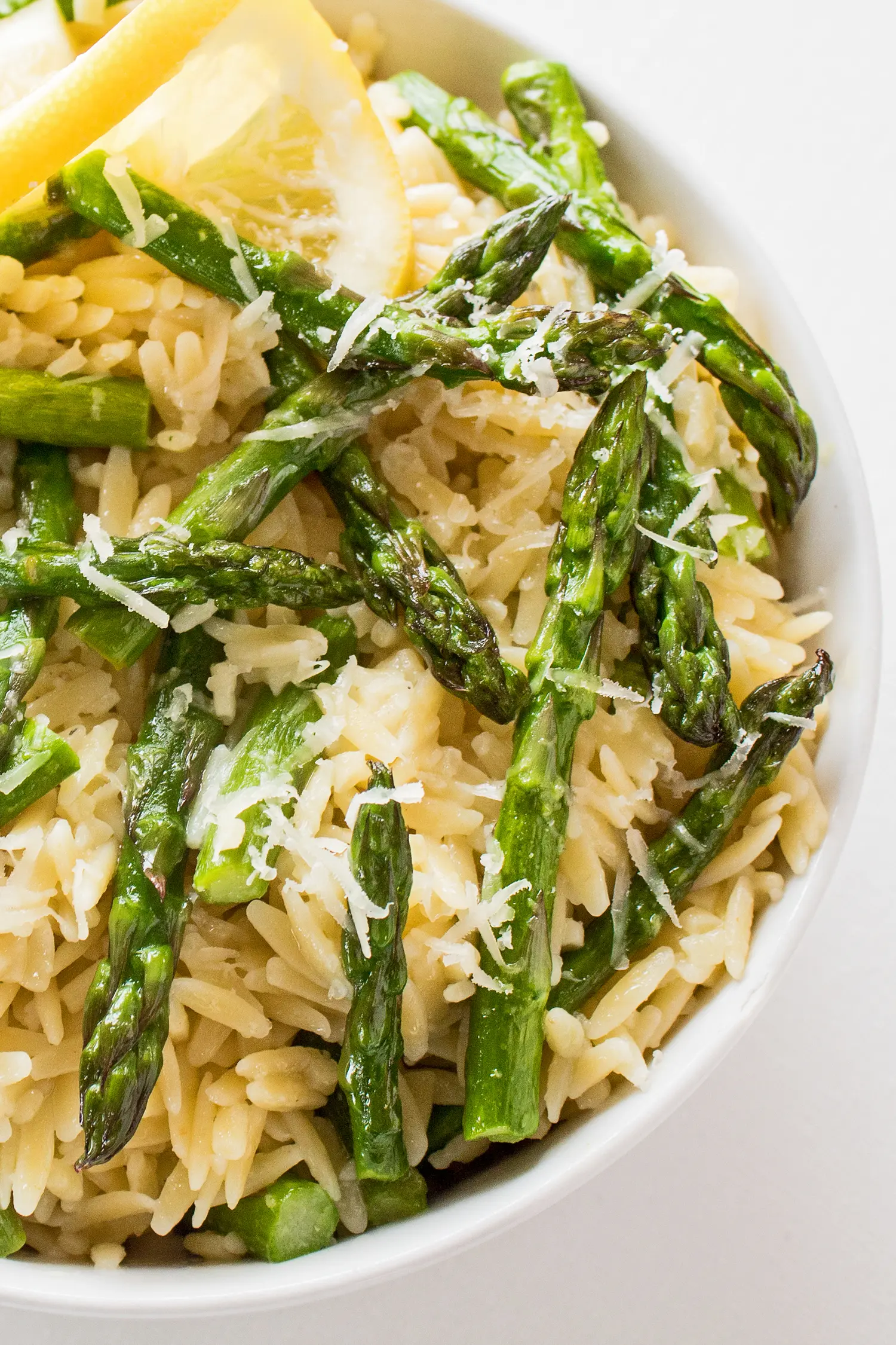 vertical overhead image of orzo pasta with asparagus and lemon sauce garnished with grated parmesan and a lemon twist in a white bowl and background