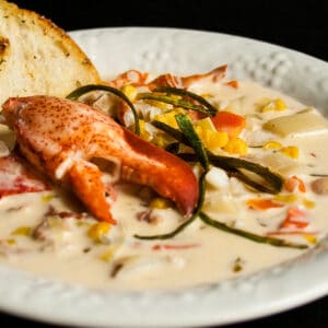 large square image of an angled overhead view of lobster chowder served in a white rimmed bowl on black background garnished with fresh cooked lobster claw fried chives roasted corn and toasted bread