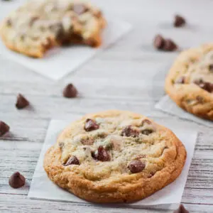 A large square image of chocolate chip cookie on parchment paper square with a light grey wooden grain background and a few scattered chocolate chips with parts of other cookies in the background.