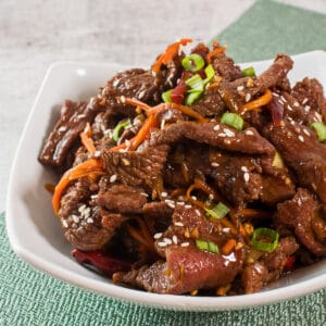 Large square image showing cooked mongolian beef from a slightly overhead side view in a white bowl on light background.