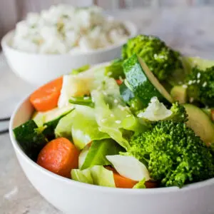 large square image taken from a side view of the panda express mixed vegetables served in a white bowl on a light background with a bowl of rice in the background