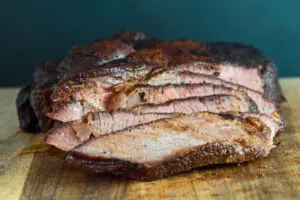 a closeup horizontal image showing tender juicy sliced smoked beef chuck roast after smoking on the cutting board with dark teal background
