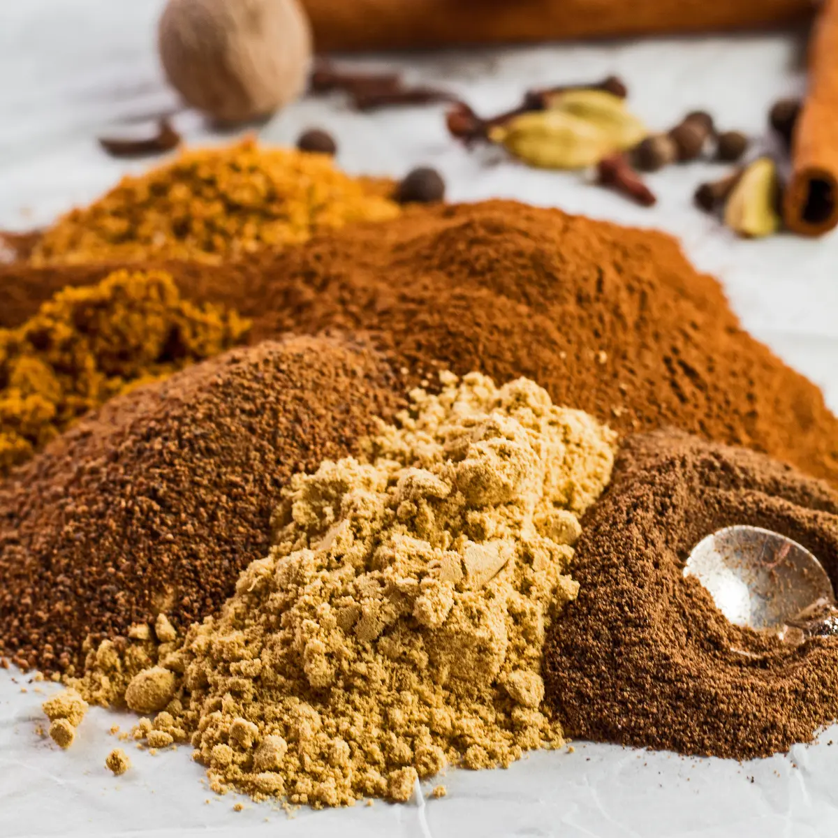 Large square image of the ground spices poured onto a white background with whole spices for the pumpkin pie spice in background.