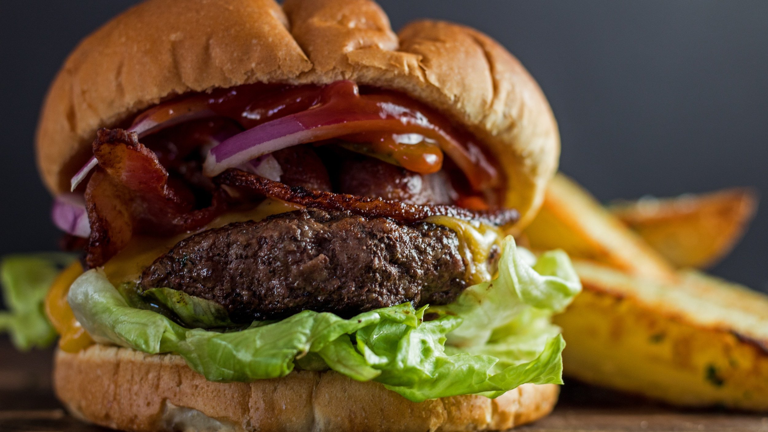 Wide image of Wagyu beef burger with fried potato wedges in background.