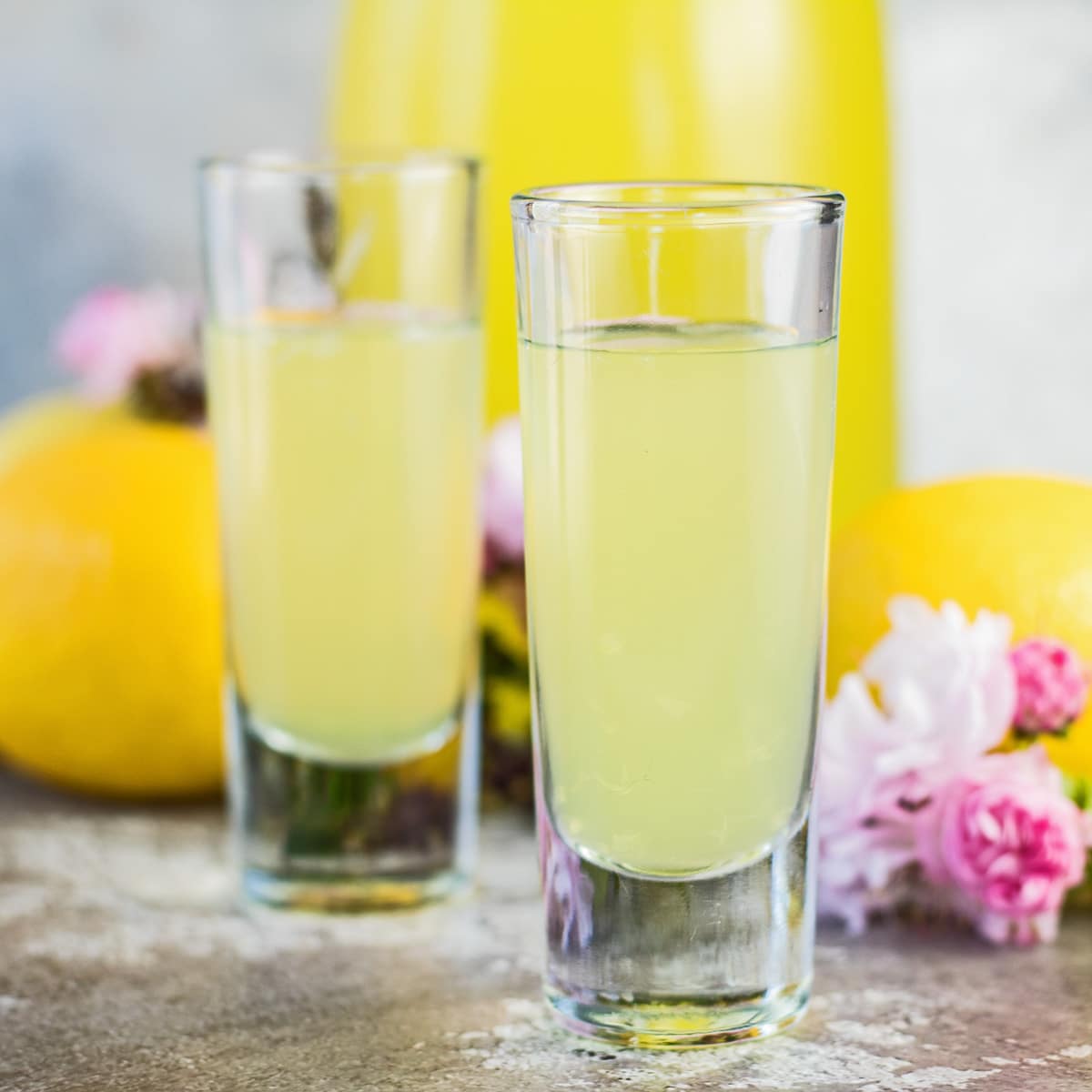 Lemoncello fatto in casa servito in bicchierini con frutta e fiori rosa in sottofondo.
